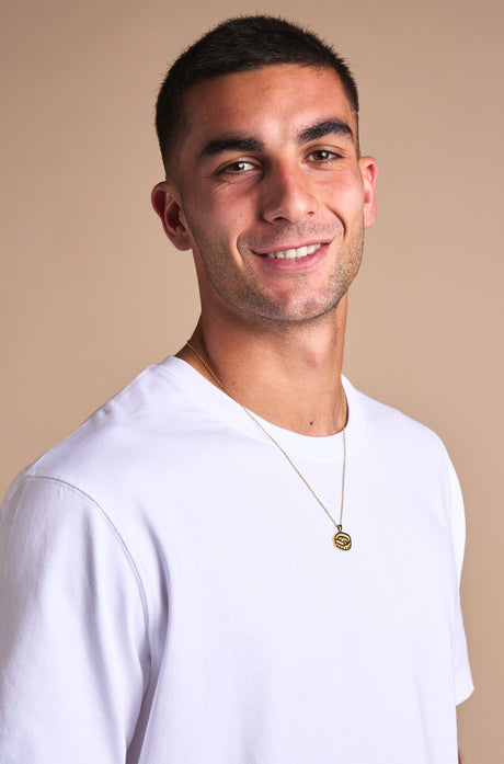 Gold necklace with a pendant in the shape of Spotify Camp Nou stadium, featuring a central sparkling gemstone, displayed between two textured rocks.