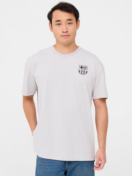 Back of a light beige T-shirt featuring a playful blue FC Barcelona crest cartoon kicking a soccer ball. Above it reads “FC BARCELONA” and below, in bold, “BLAU GRANA SÓN ELS COLORS” with “EST. 1899” on each side