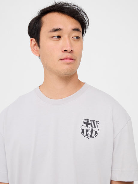 Back of a light beige T-shirt featuring a playful blue FC Barcelona crest cartoon kicking a soccer ball. Above it reads “FC BARCELONA” and below, in bold, “BLAU GRANA SÓN ELS COLORS” with “EST. 1899” on each side