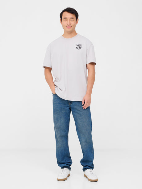 Back of a light beige T-shirt featuring a playful blue FC Barcelona crest cartoon kicking a soccer ball. Above it reads “FC BARCELONA” and below, in bold, “BLAU GRANA SÓN ELS COLORS” with “EST. 1899” on each side