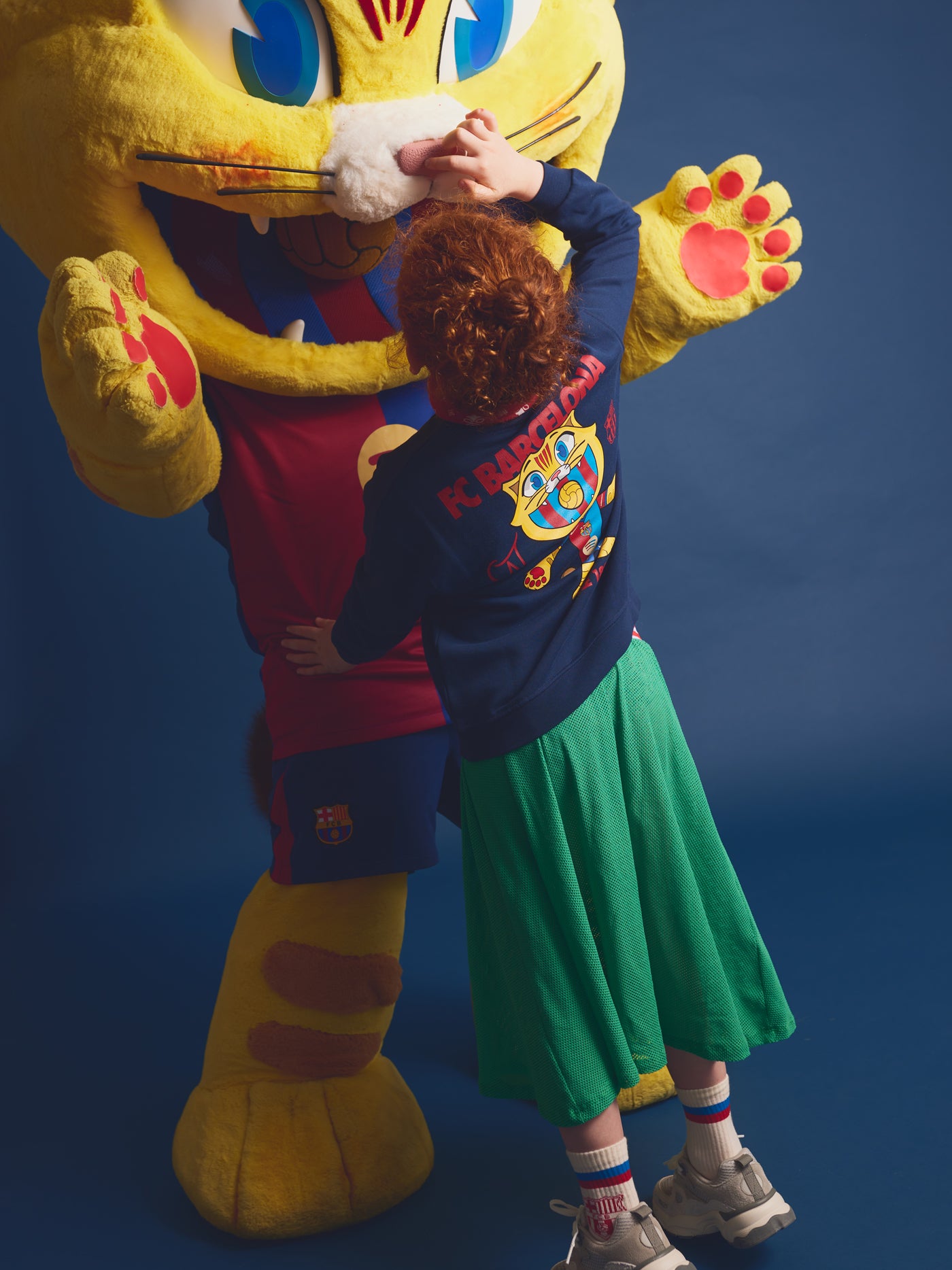 Girl hugging Barça’s mascot, Cat Culer, wearing a navy blue sweatshirt with a colorful Cat Culer illustration on the back, green skirt, and Barça socks.