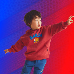 Child wearing a red FC Barcelona hoodie with the club crest and blue lettering, paired with blue pants, posing playfully against a red and blue gradient background