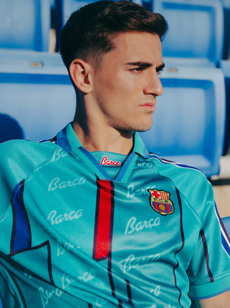 Teal short-sleeve polo shirt with FC Barcelona crest, blue and red accents, and Barça-themed script print.