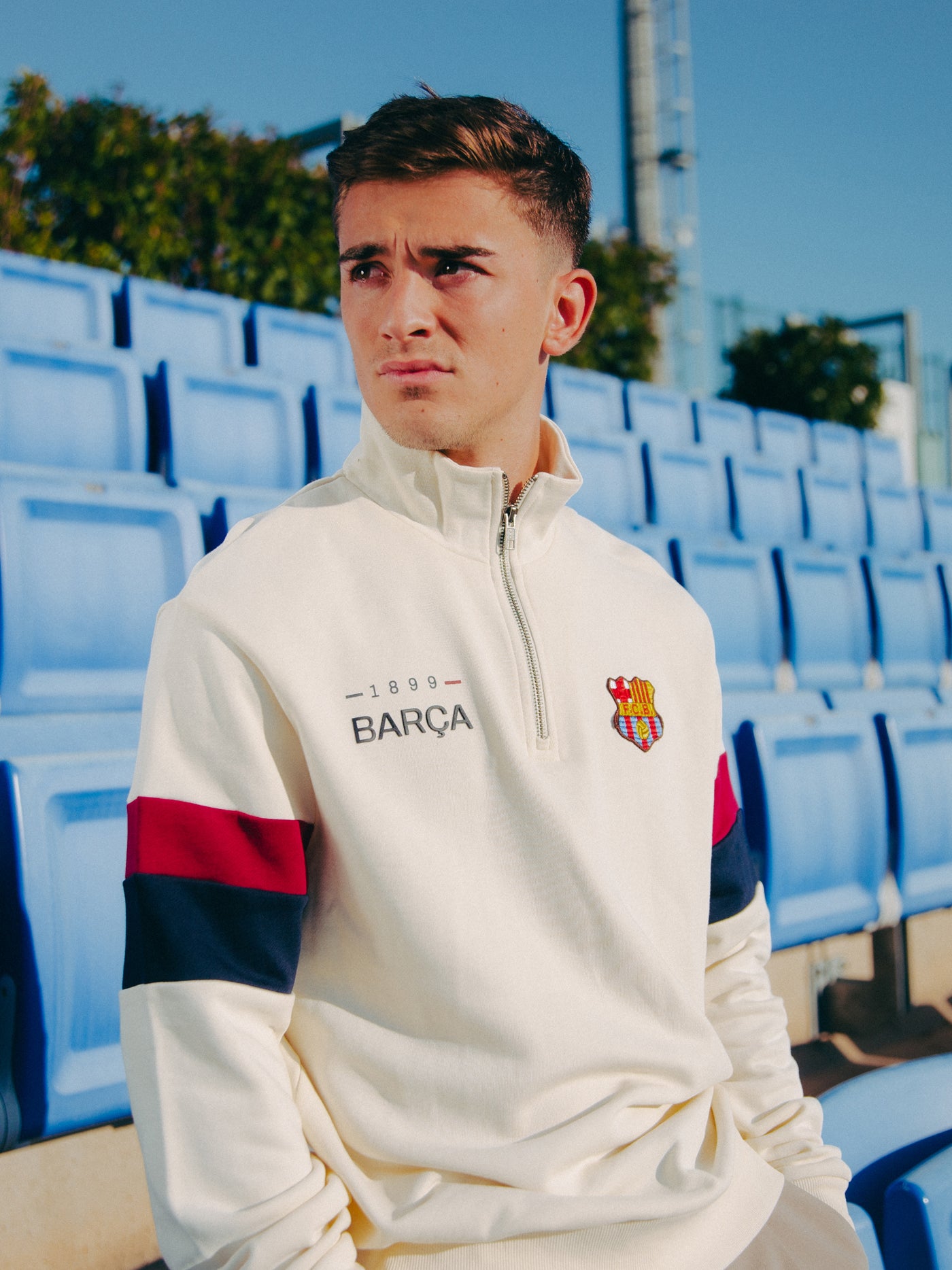 Close-up of Gavi seated in a stadium wearing a cream-colored Barça sweatshirt with red and navy sleeve stripes and embroidered club crest, evoking a calm and stylish atmosphere.