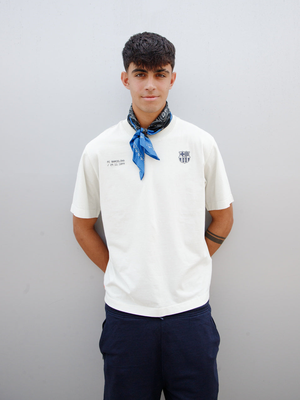 Marc Bernal wearing an off-white FC Barcelona T-shirt with tonal club crest and date detail on the chest, paired with a blue and black patterned neck scarf