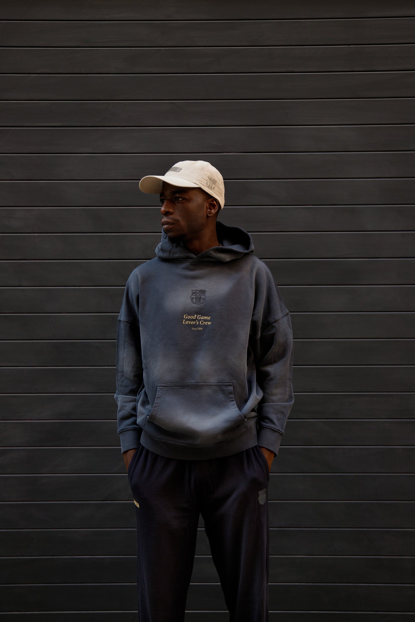 Man wearing a dark blue FC Barcelona hoodie with subtle crest and “Good Game Lover’s Crew” text on the chest, paired with matching joggers and a beige cap, standing against a dark background.
