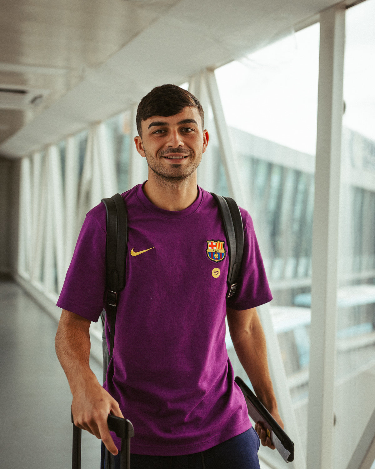 Pedri wearing a purple FC Barcelona T-shirt with the club crest and Nike logo, standing indoors with a backpack and suitcase.