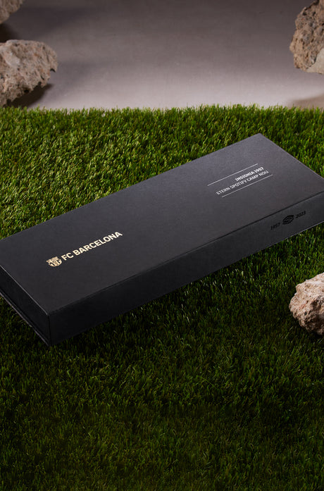 Gold necklace with a pendant in the shape of Spotify Camp Nou stadium, featuring a central sparkling gemstone, displayed between two textured rocks.