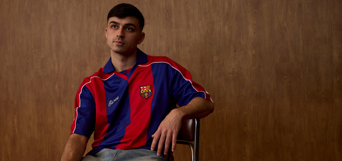 Front view of an FC Barcelona jersey with blue and red vertical stripes, shown on a seated model with the club crest on the chest.