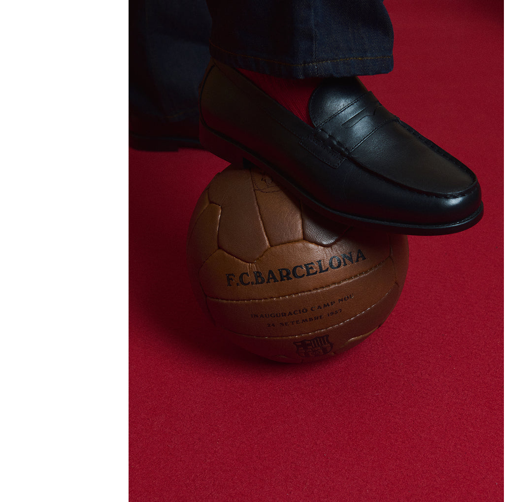 Black leather loafer resting on a vintage F.C. Barcelona soccer ball against a red background.