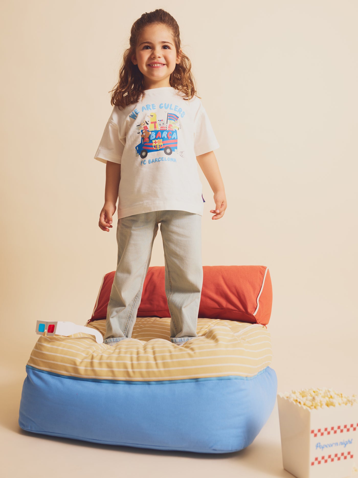 Child smiling and standing on a cushion, wearing a white FC Barcelona T-shirt with colorful cartoon illustrations and the text 'We Are Culers
