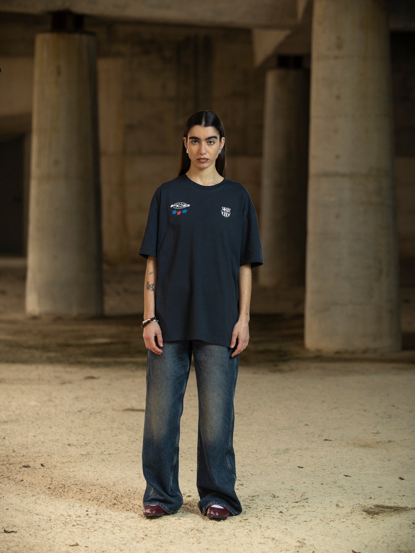 Person wearing a dark blue t-shirt and jeans standing in an industrial setting with concrete pillars.