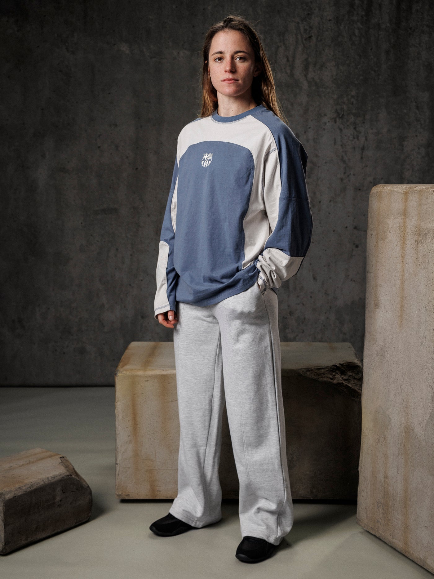 Person wearing a blue and white long-sleeve shirt with a logo, standing against a textured gray wall.