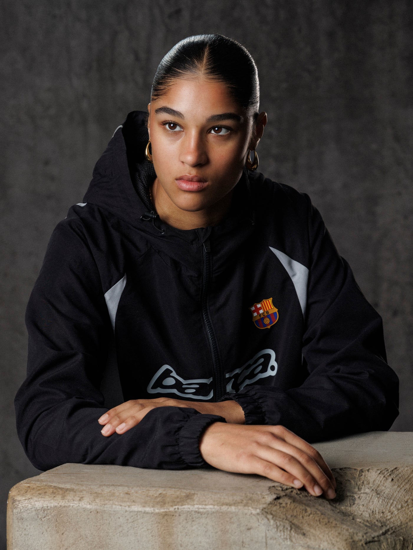 Person wearing a black jacket with a logo, sitting on a stone surface against a dark background