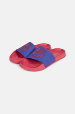 A pair of red and blue FC Barcelona slide sandals held up against a clear blue sky. The sandals feature the FC Barcelona crest prominently on the blue strap across the top.