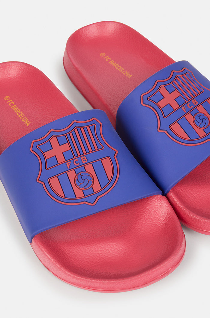 A pair of red and blue FC Barcelona slide sandals held up against a clear blue sky. The sandals feature the FC Barcelona crest prominently on the blue strap across the top.