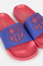 A pair of red and blue FC Barcelona slide sandals held up against a clear blue sky. The sandals feature the FC Barcelona crest prominently on the blue strap across the top.