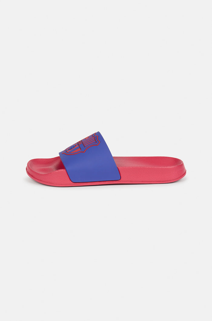 A pair of red and blue FC Barcelona slide sandals held up against a clear blue sky. The sandals feature the FC Barcelona crest prominently on the blue strap across the top.