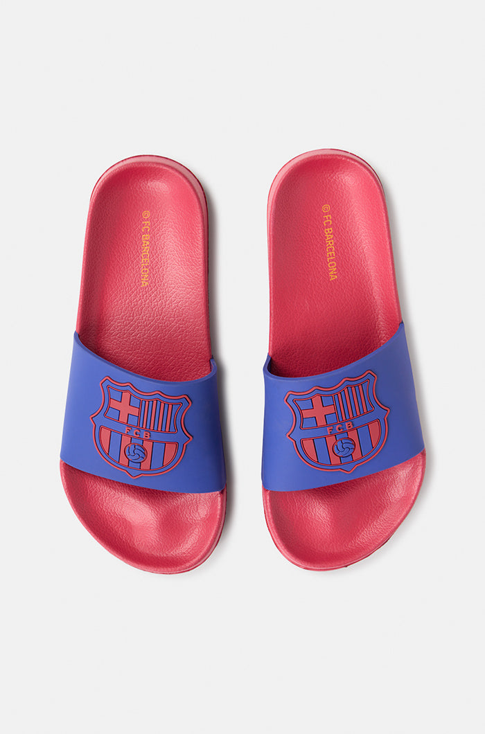 A pair of red and blue FC Barcelona slide sandals held up against a clear blue sky. The sandals feature the FC Barcelona crest prominently on the blue strap across the top.