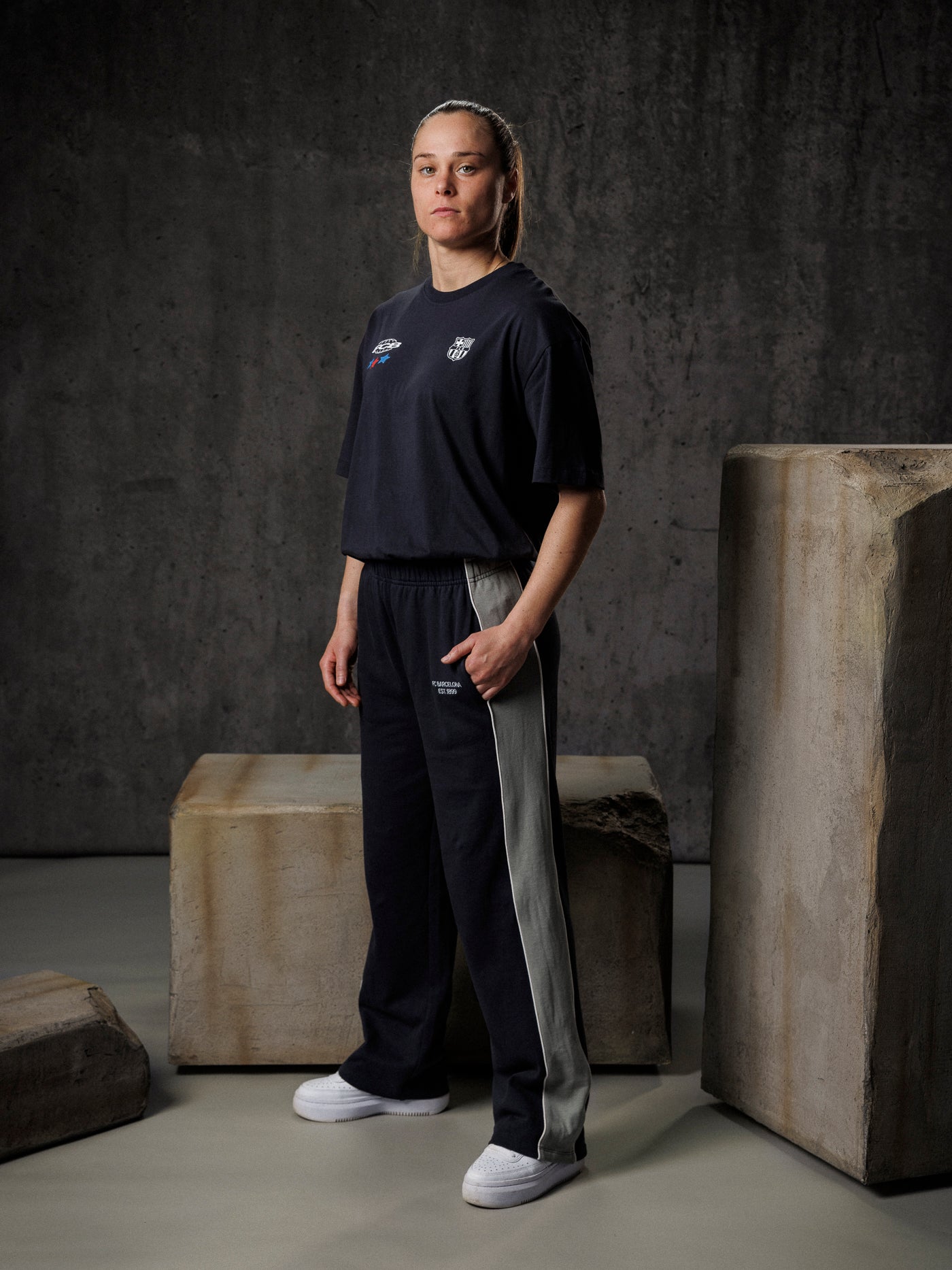 Person wearing a black t-shirt and pants standing against a dark background with stone blocks.