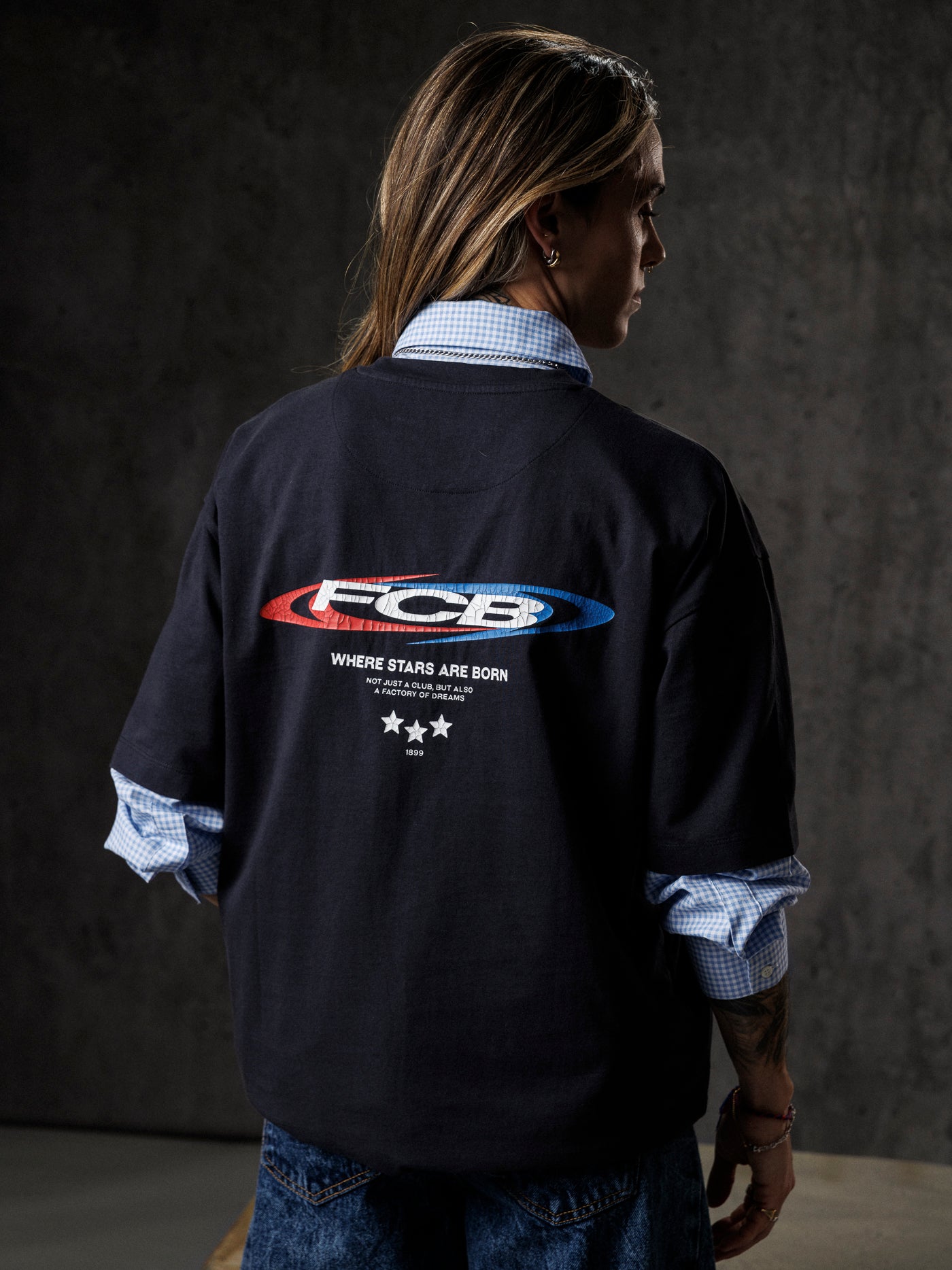 Person wearing a black jacket with 'FCB' logo on the back against a dark background