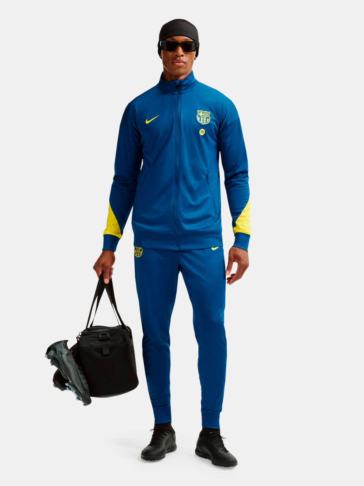 Person wearing a blue sports tracksuit with yellow accents, holding soccer cleats and a bag on a white background.