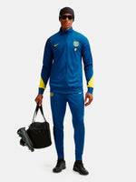 Person wearing a blue sports tracksuit with yellow accents, holding soccer cleats and a bag on a white background.
