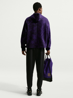 Vista trasera de un modelo con sudadera con capucha FC Barcelona de efecto mármol morado, pantalón negro y bolsa con cordón morada con el escudo del Barça.