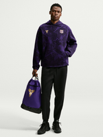 Modelo con sudadera con capucha morada de FC Barcelona con escudo dorado y logo de Kobe, pantalón negro y bolsa morada con cordón a juego.