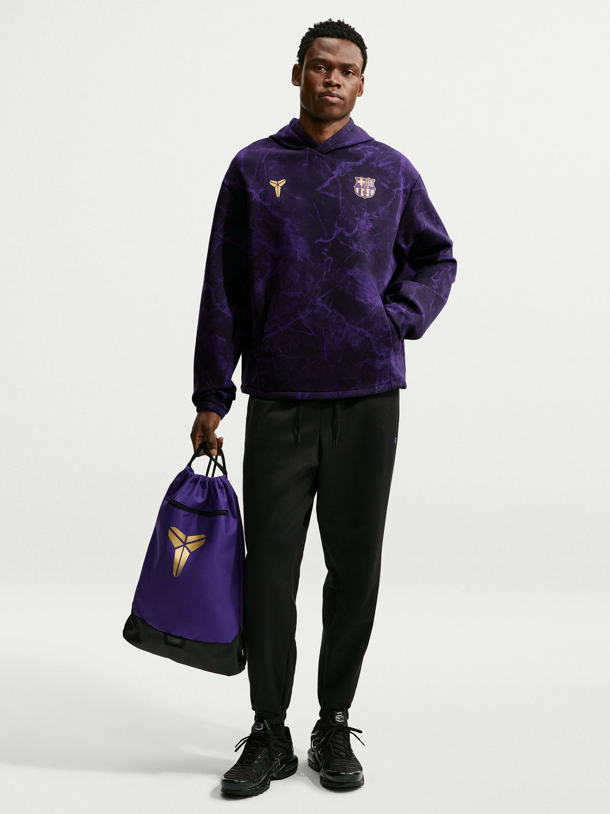 Modelo con sudadera con capucha morada de FC Barcelona con escudo dorado y logo de Kobe, pantalón negro y bolsa morada con cordón a juego.