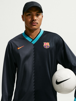 Vista frontal de una camiseta negra de manga larga de FC Barcelona con ribete turquesa en cuello en V, logo de Nike, escudo del club y un balón blanco de Nike sujeto al lado.