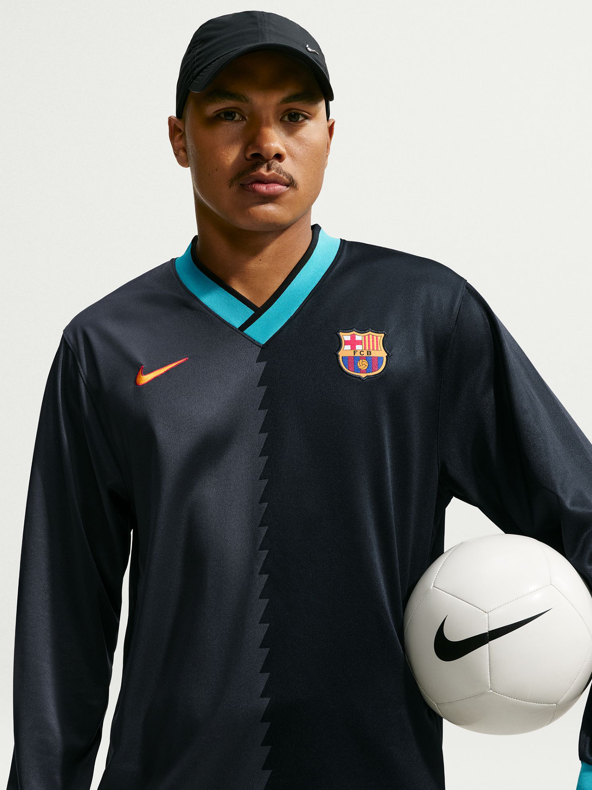 Vista frontal de una camiseta negra de manga larga de FC Barcelona con ribete turquesa en cuello en V, logo de Nike, escudo del club y un balón blanco de Nike sujeto al lado.