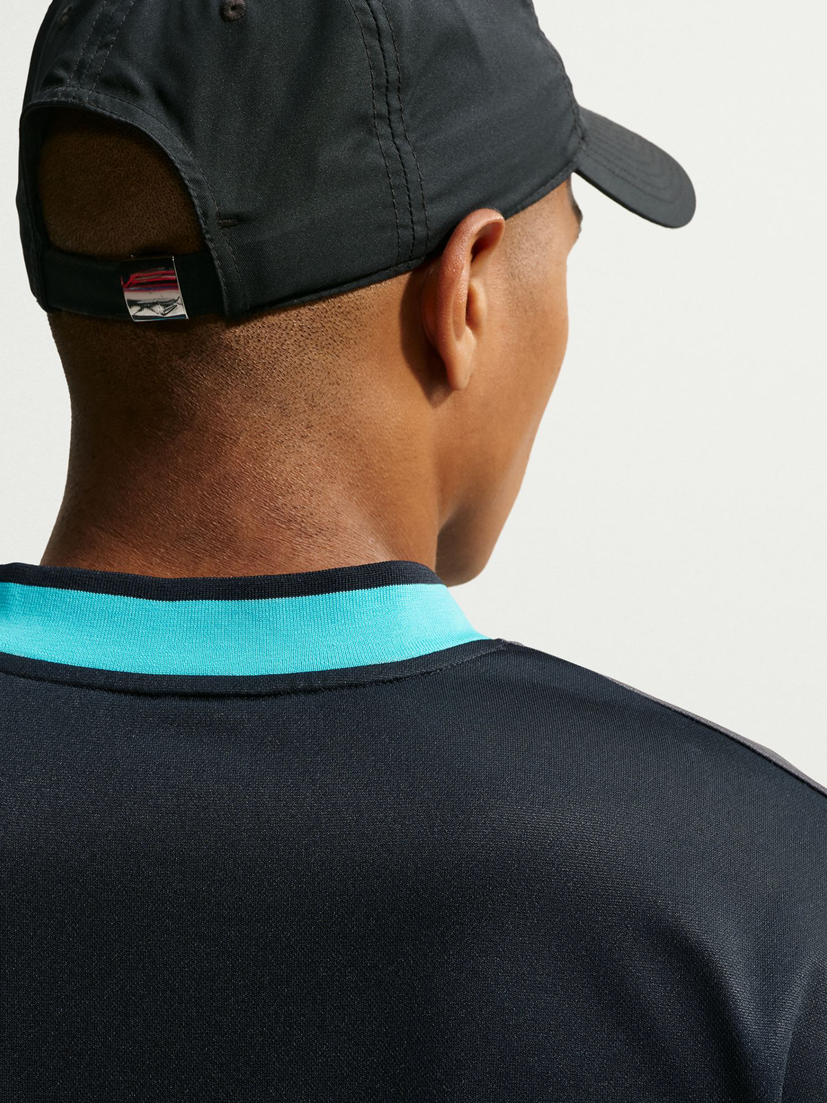 Vista trasera de un modelo con gorra negra y camiseta negra del FC Barcelona con cuello turquesa.