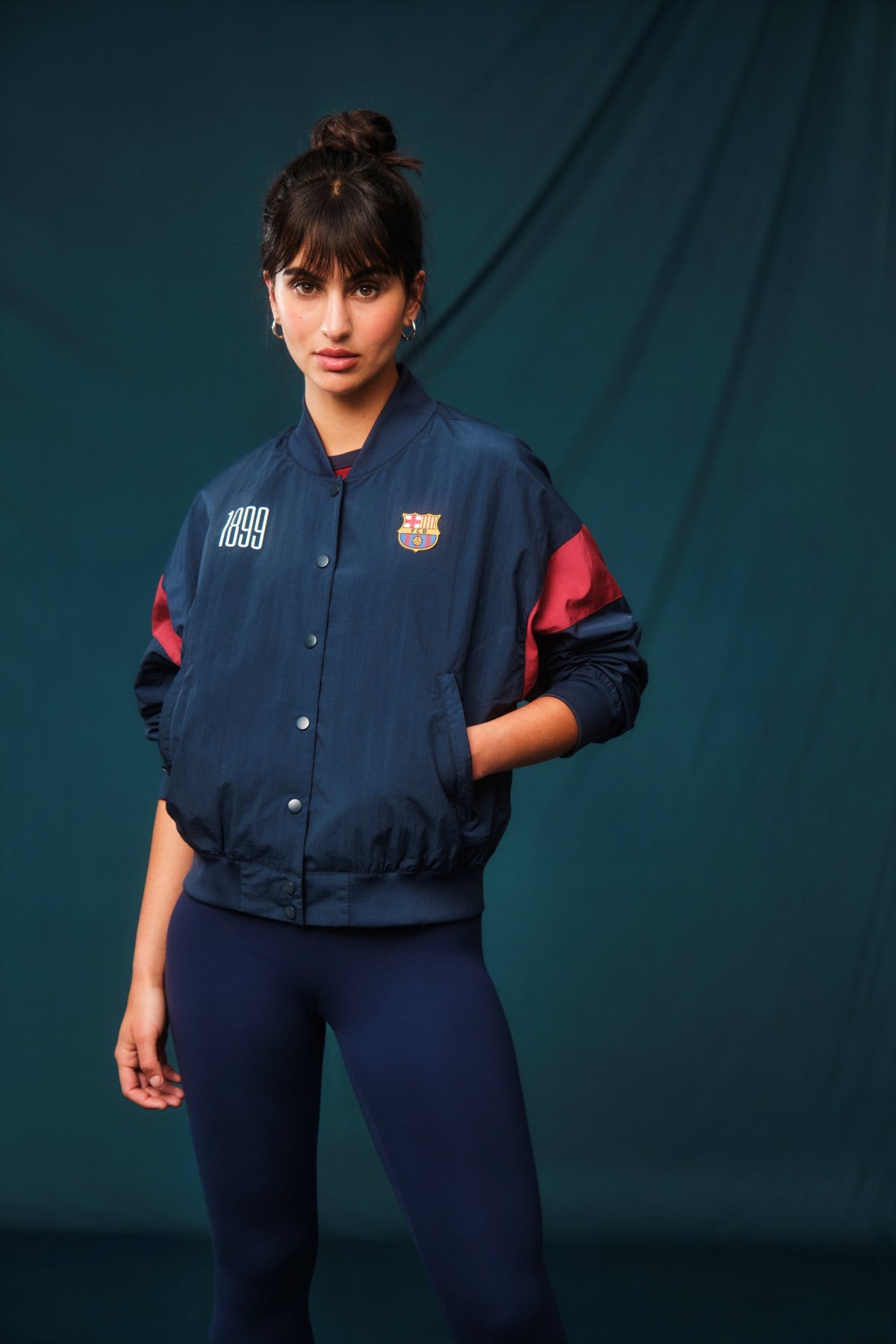 Navy blue FC Barcelona bomber jacket with red bands on the sleeves. The jacket features the club crest on the left chest and the year "1899" on the right chest, with a front snap button closure and ribbed cuffs and hem.