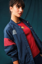 Navy blue FC Barcelona bomber jacket with red bands on the sleeves. The jacket features the club crest on the left chest and the year "1899" on the right chest, with a front snap button closure and ribbed cuffs and hem.
