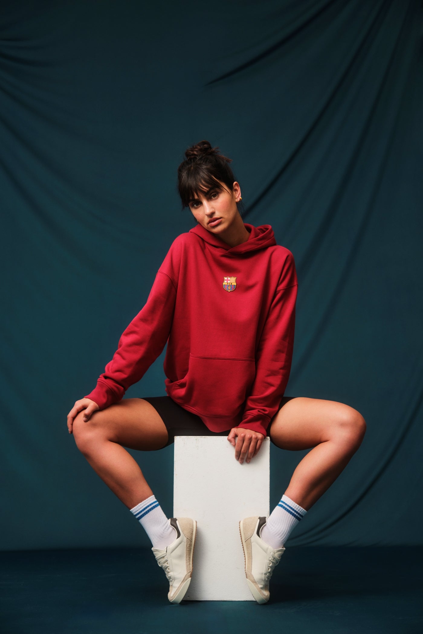 Woman wearing a red FC Barcelona hoodie featuring the club’s crest on the chest. She is adjusting the hood with both hands while slightly turning her head to the side, showing off gold hoop earrings.








