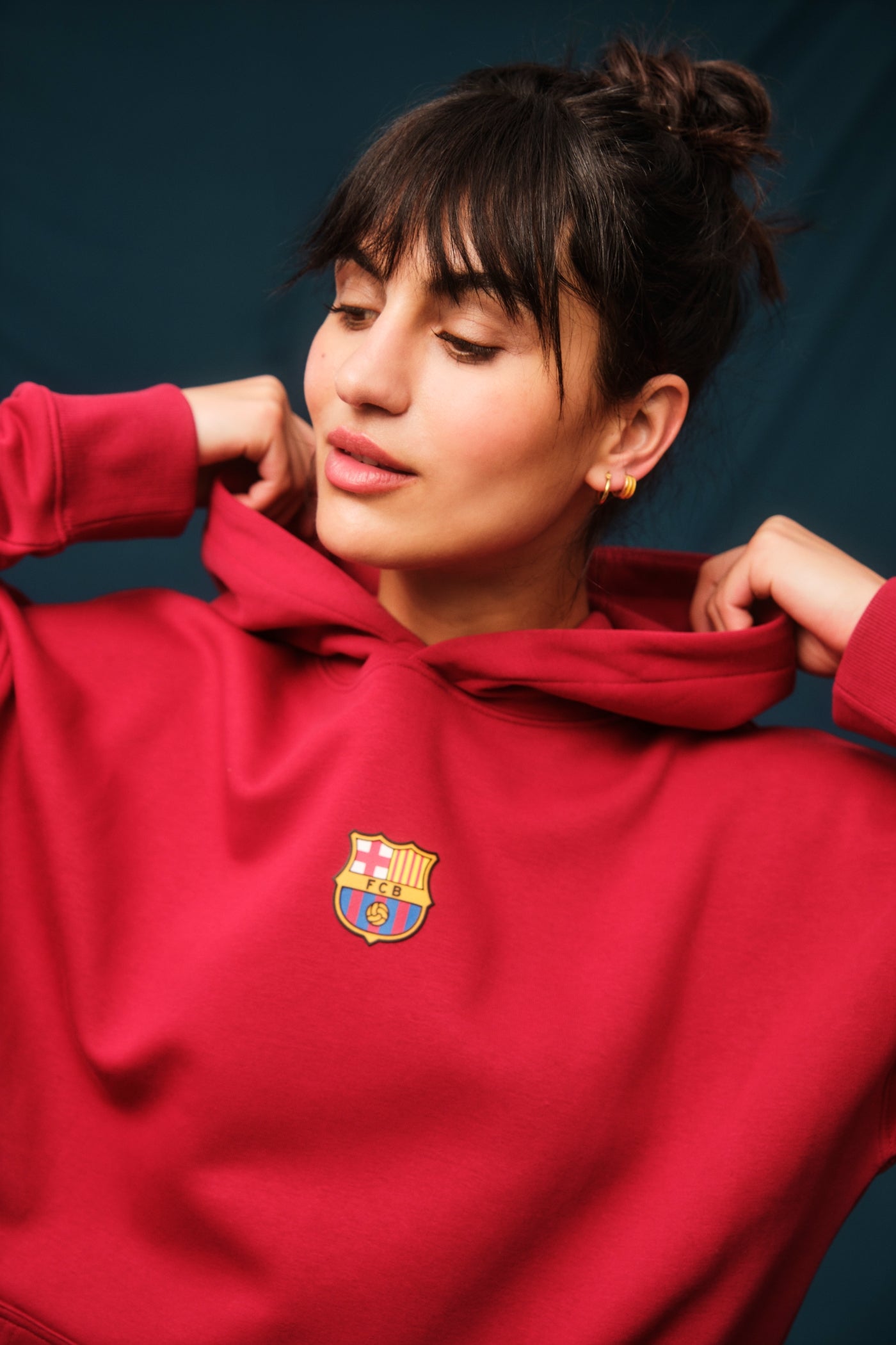 Woman wearing a red FC Barcelona hoodie featuring the club’s crest on the chest. She is adjusting the hood with both hands while slightly turning her head to the side, showing off gold hoop earrings.