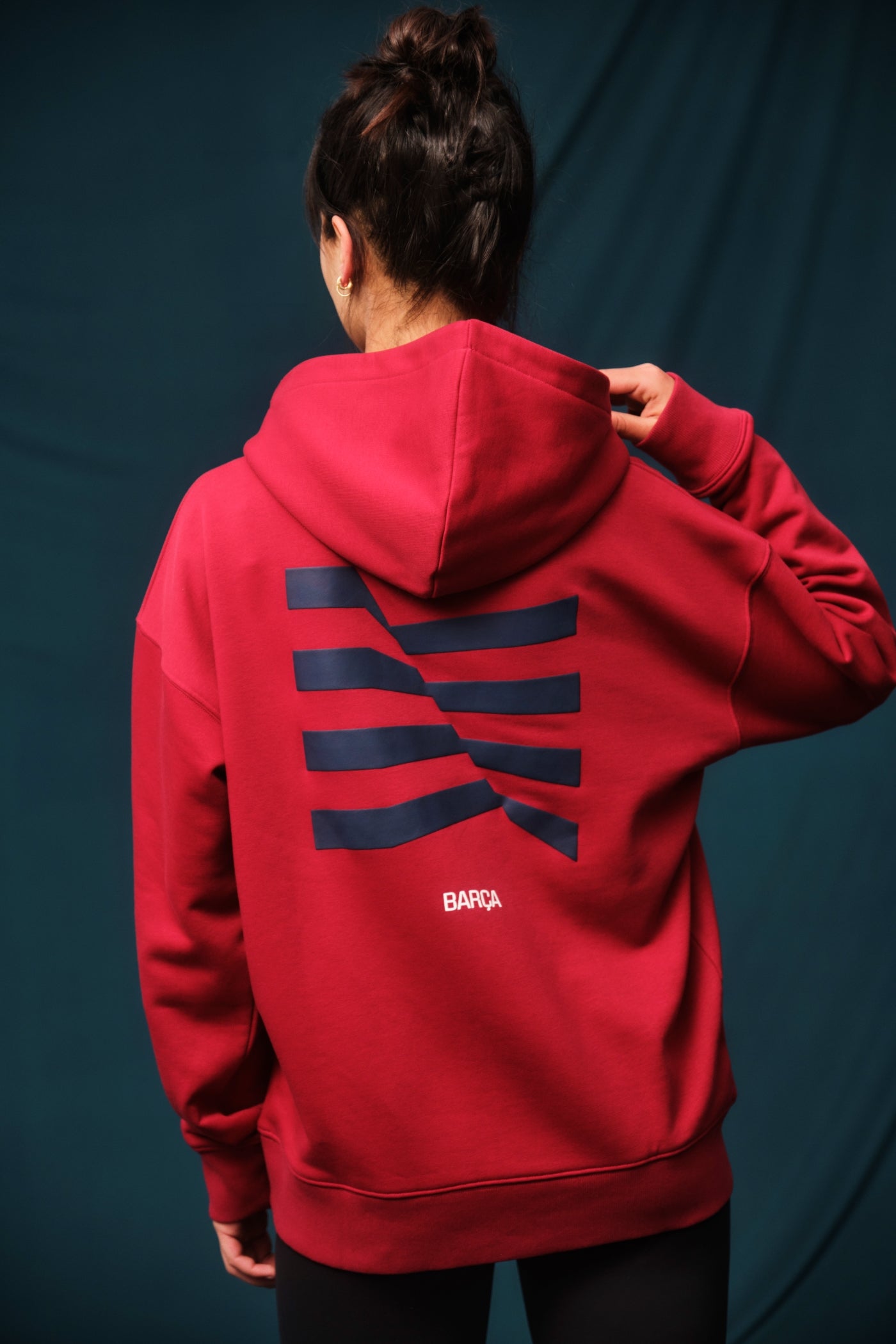 Woman wearing a red FC Barcelona hoodie featuring the club’s crest on the chest. She is adjusting the hood with both hands while slightly turning her head to the side, showing off gold hoop earrings.








