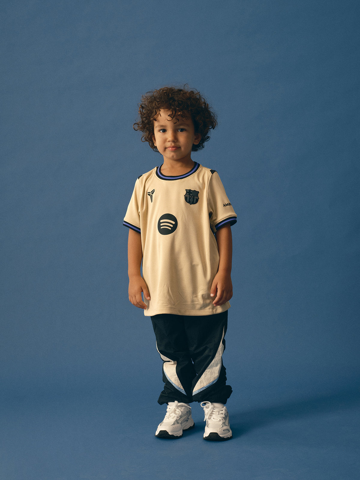 FC Barcelona kids' away kit set including beige jersey with black and purple details, black shorts with purple accents, and matching black socks, featuring club crest, Nike logo, Spotify sponsor, and Kobe Bryant tribute emblem.
