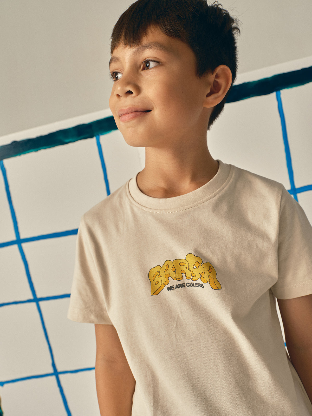 Beige FC Barcelona T-shirt featuring a vibrant vintage-style back print with the word "Barça" in large retro bubble letters. The graphic includes a football, flags, and stadium elements in bold colors within a shield-like frame. A ribbon below the design reads “Since 1899” and “Més que un club.”