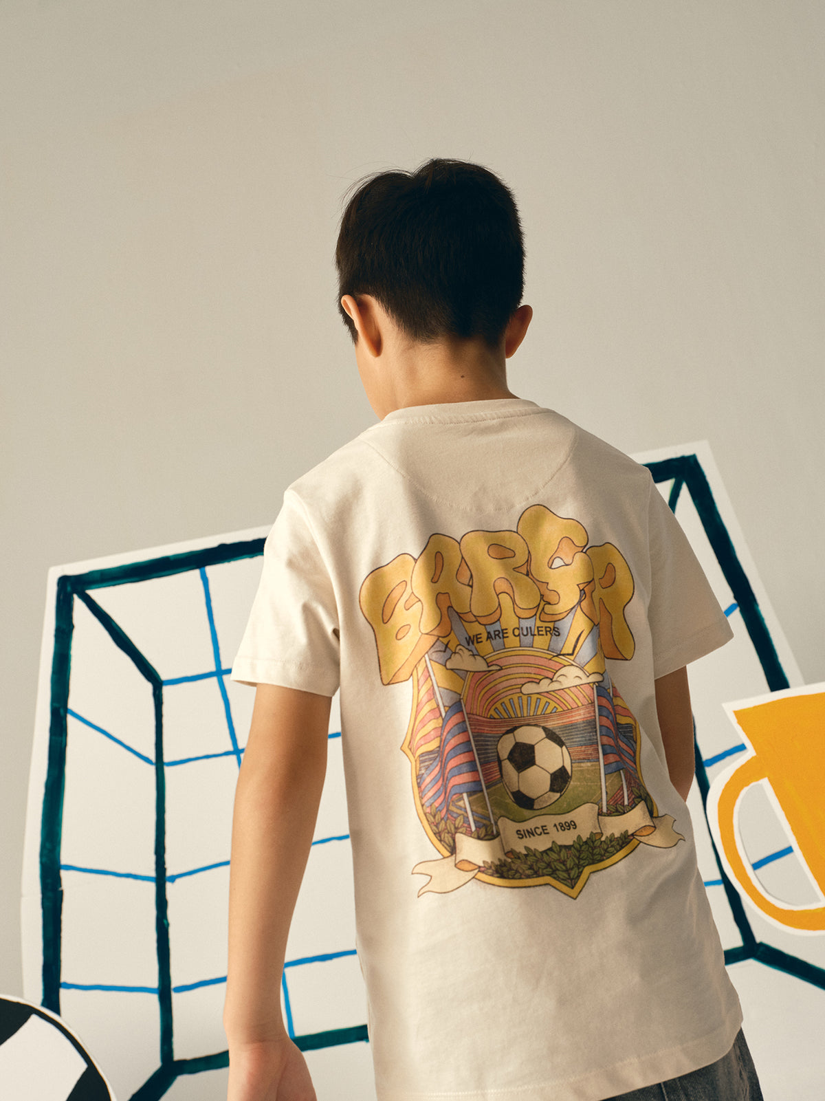 Beige FC Barcelona T-shirt featuring a vibrant vintage-style back print with the word "Barça" in large retro bubble letters. The graphic includes a football, flags, and stadium elements in bold colors within a shield-like frame. A ribbon below the design reads “Since 1899” and “Més que un club.”