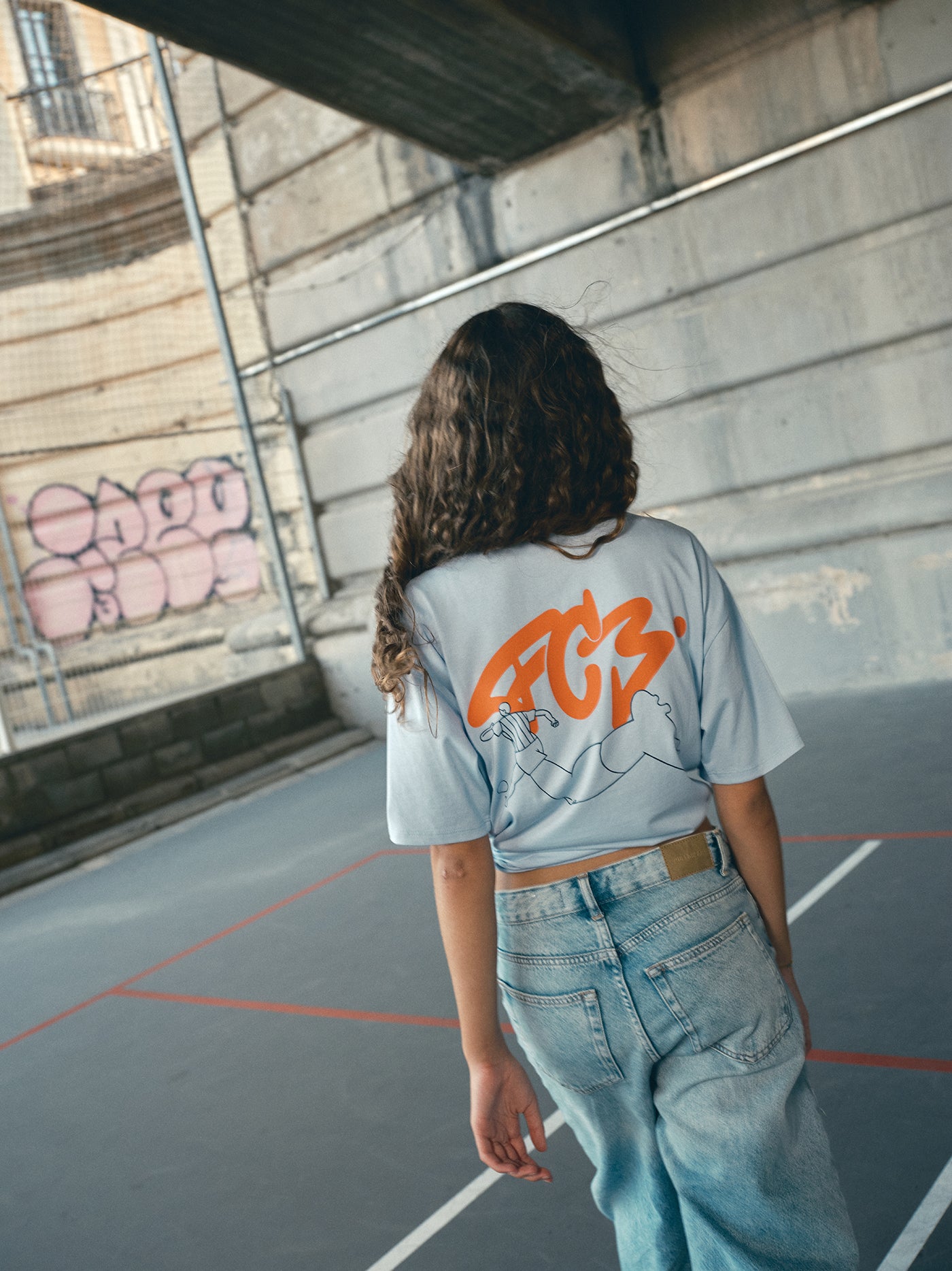 light blue FC Barcelona t-shirt with a large orange 'FCB' graphic and a black line illustration of a footballer kicking a ball