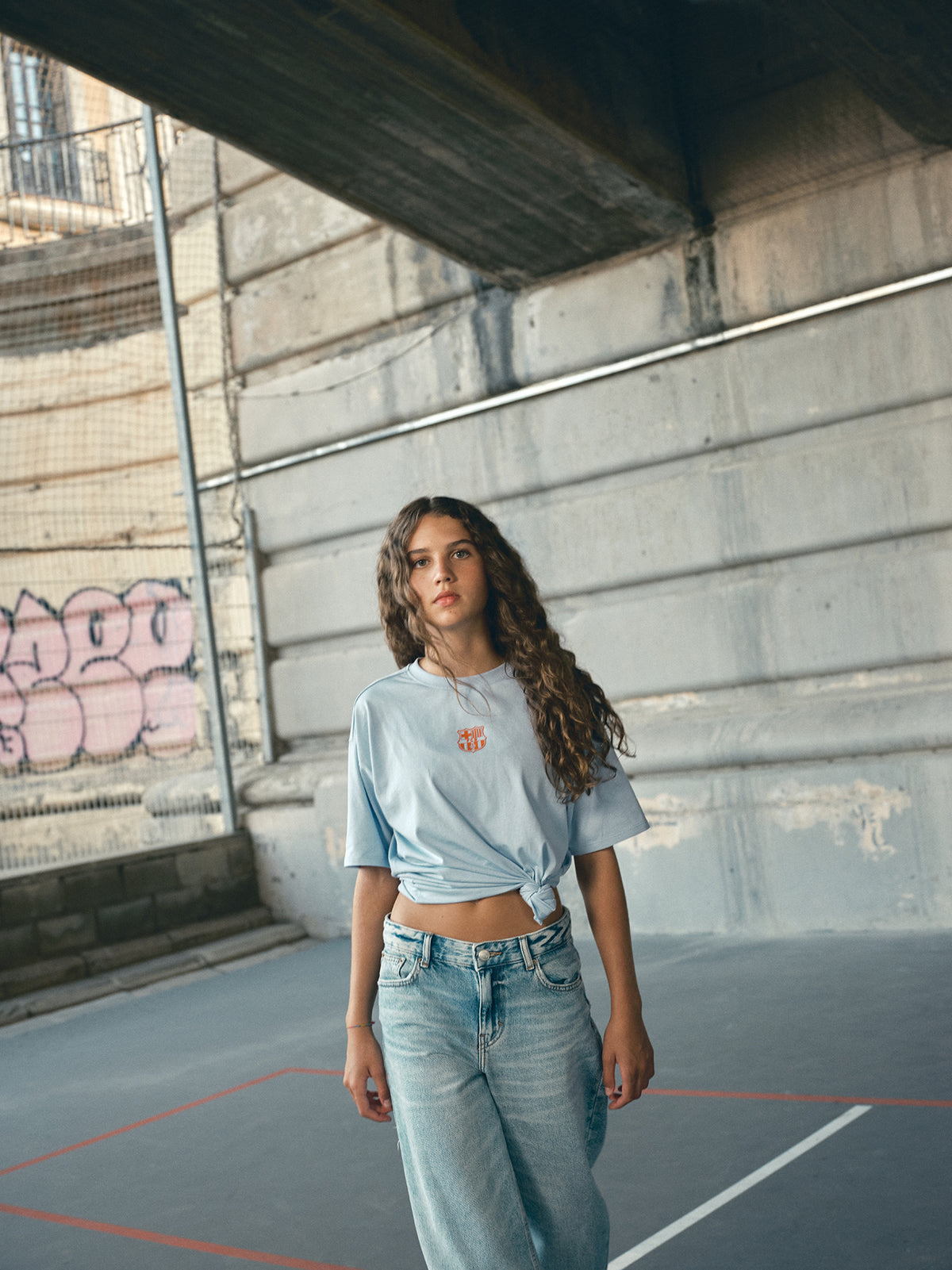 light blue FC Barcelona t-shirt with a large orange 'FCB' graphic and a black line illustration of a footballer kicking a ball
