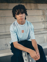 light blue FC Barcelona t-shirt with a large orange 'FCB' graphic and a black line illustration of a footballer kicking a ball