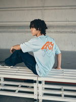 light blue FC Barcelona t-shirt with a large orange 'FCB' graphic and a black line illustration of a footballer kicking a ball