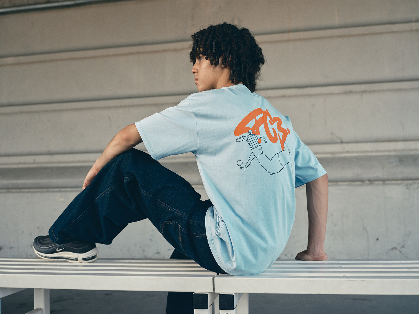 light blue FC Barcelona t-shirt with a large orange 'FCB' graphic and a black line illustration of a footballer kicking a ball
