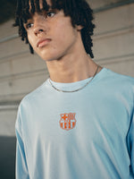 light blue FC Barcelona t-shirt with a large orange 'FCB' graphic and a black line illustration of a footballer kicking a ball
