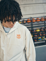 Beige FC Barcelona hooded jacket with minimalist orange graphic icons and text printed across the upper back.