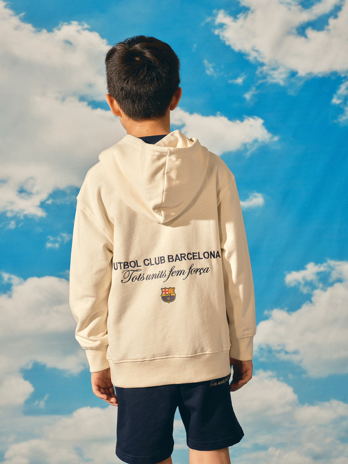 Beige FC Barcelona hoodie for kids, featuring a colorful 'Barça' text on the front and 'Futbol Club Barcelona - Tots units fem força' with the club crest on the back