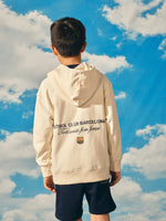 Beige FC Barcelona hoodie for kids, featuring a colorful 'Barça' text on the front and 'Futbol Club Barcelona - Tots units fem força' with the club crest on the back
